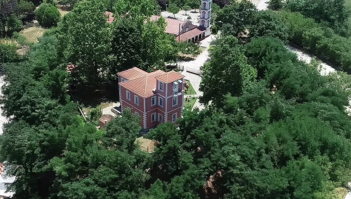 κόκκινο κτίριο του Αρχαιολογικού Μουσείου Καρδίτσας, περιτριγυρισμένο από πυκνή, καταπράσινη βλάστηση και ψηλά δέντρα. Στο βάθος διακρίνεται μια εκκλησία με καμπαναριό.