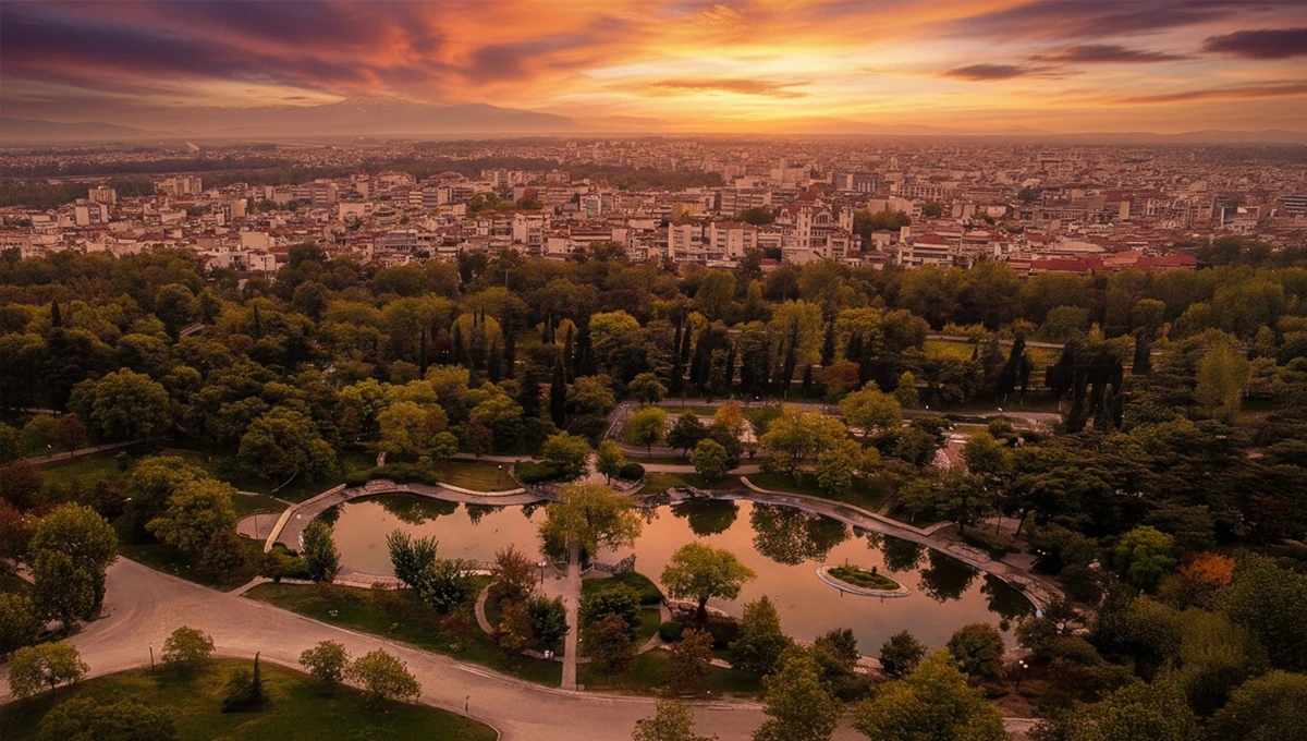 Αεροφωτογραφία του Πάρκου του Αλκαζάρ στη Λάρισα με την τεχνητή λίμνη και πλούσια βλάστηση, την πόλη στο βάθος και εντυπωσιακό ηλιοβασίλεμα.