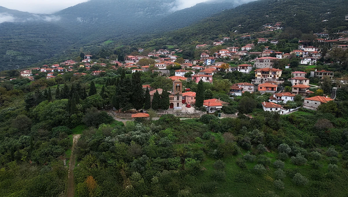 Αεροφωτογραφία του ιστορικού χωριού Αμπελάκια, χτισμένο αμφιθεατρικά σε καταπράσινη πλαγιά με παραδοσιακά σπίτια και μια εκκλησία στο κέντρο.