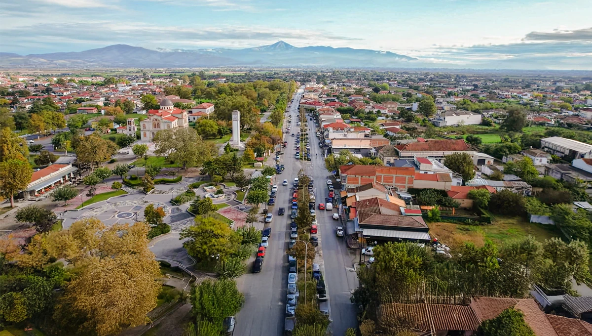 Αεροφωτογραφία του Αμπελώνα Λάρισας με την κεντρική λεωφόρο, πάρκο, εκκλησία και κατοικίες, με φόντο βουνά.