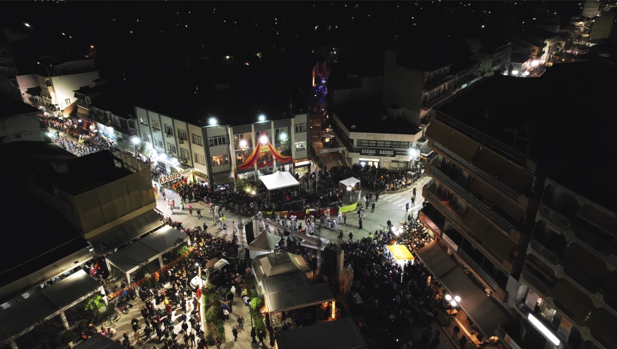 Νυχτερινή αεροφωτογραφία μιας πλατείας στον Τύρναβο, γεμάτη με πλήθος κόσμου, φώτα και σκηνές, κατά τη διάρκεια αποκριάτικης εκδήλωσης ή του Καρναβαλιού.
