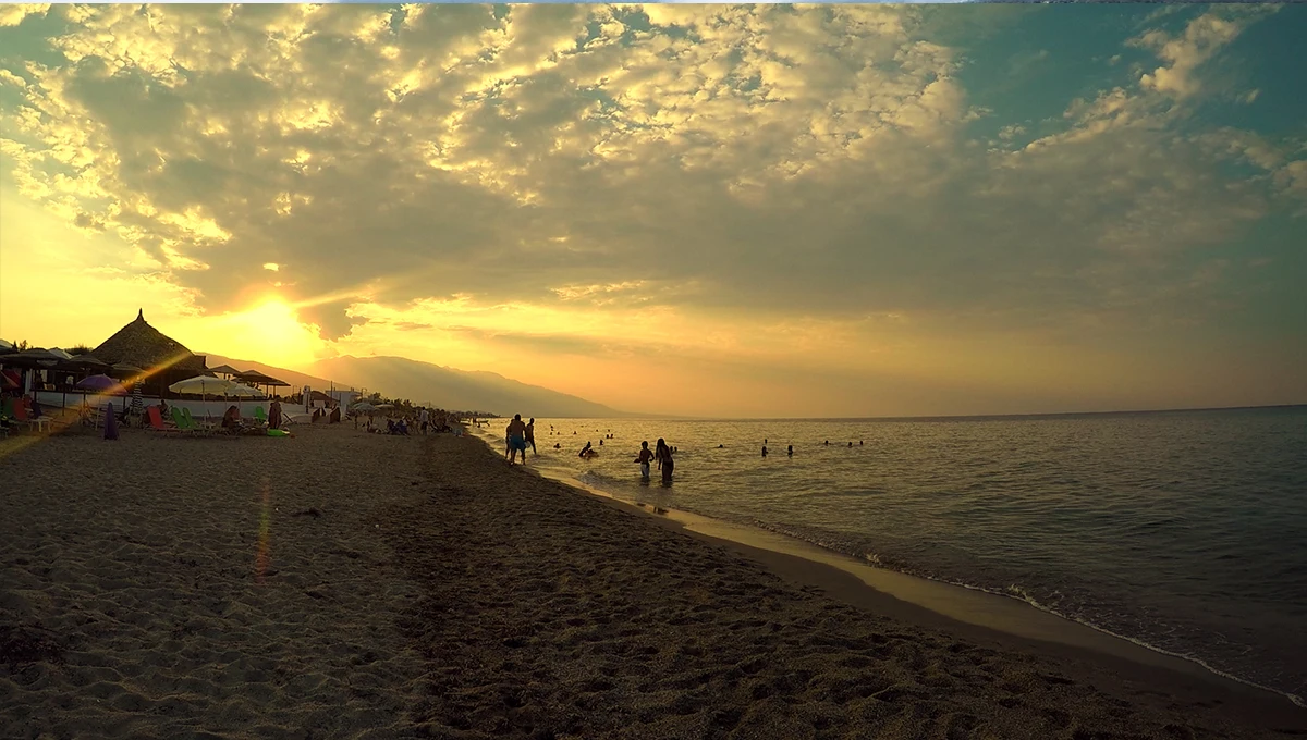 Δραματικό ηλιοβασίλεμα με χρυσαφένια σύννεφα πάνω από την Παραλία Μεσαγγάλων, με ανθρώπους να κολυμπούν και να περπατούν στην άμμο.
