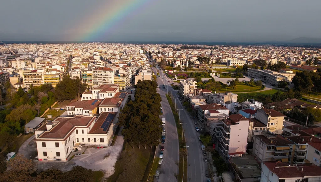 Πανοραμική αεροφωτογραφία της συνοικίας Φιλιππούπολης στη Λάρισα, με ένα φωτεινό ουράνιο τόξο στον συννεφιασμένο ουρανό πάνω από την πόλη και κεντρικούς δρόμους με δέντρα.