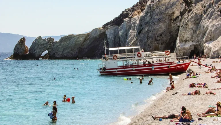 Κόσμος που κολυμπά και κάνει ηλιοθεραπεία στα λευκά βότσαλα της παραλίας Λαλάρια στη Σκιάθο, με ένα κόκκινο τουριστικό σκάφος αραγμένο και την Τρύπια Πέτρα στο βάθος.