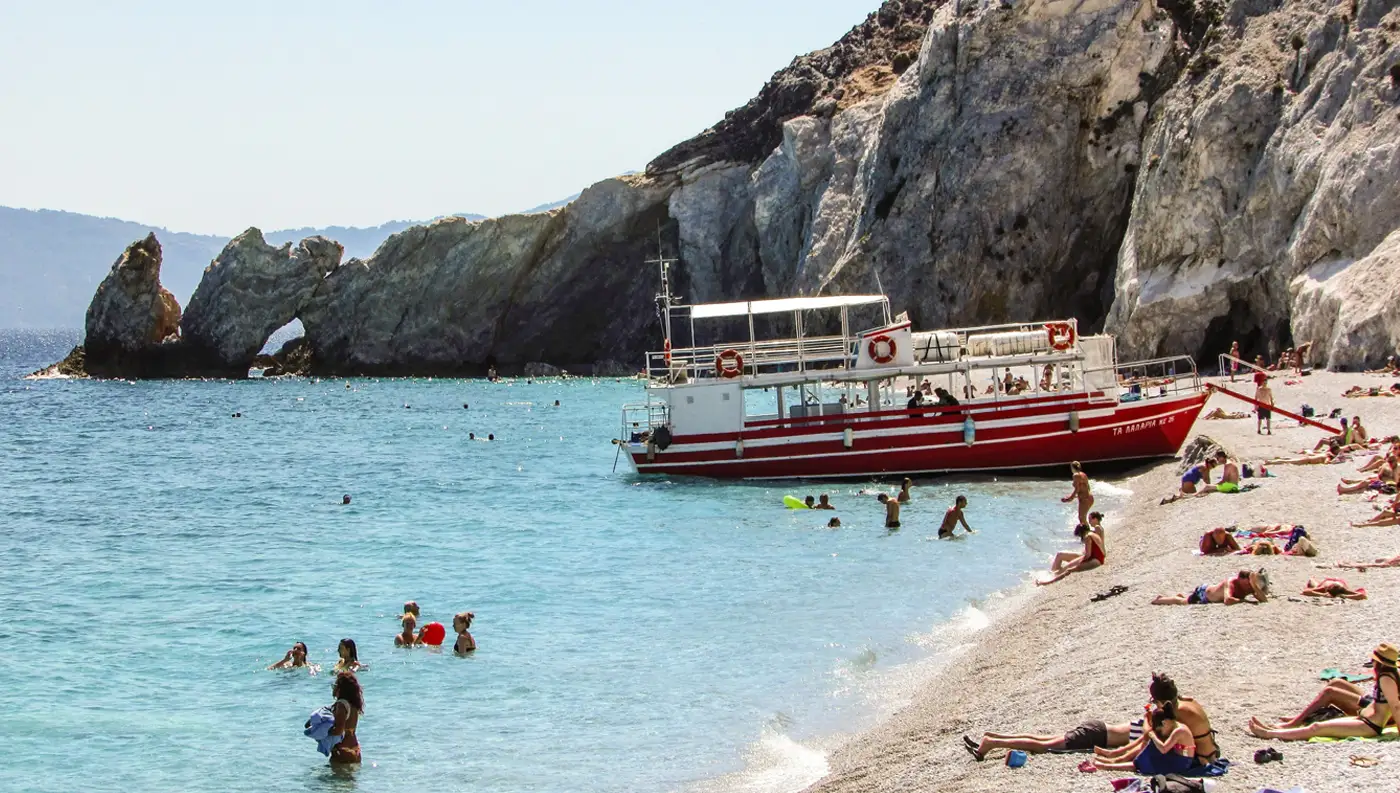 Κόσμος που κολυμπά και κάνει ηλιοθεραπεία στα λευκά βότσαλα της παραλίας Λαλάρια στη Σκιάθο, με ένα κόκκινο τουριστικό σκάφος αραγμένο και την Τρύπια Πέτρα στο βάθος.