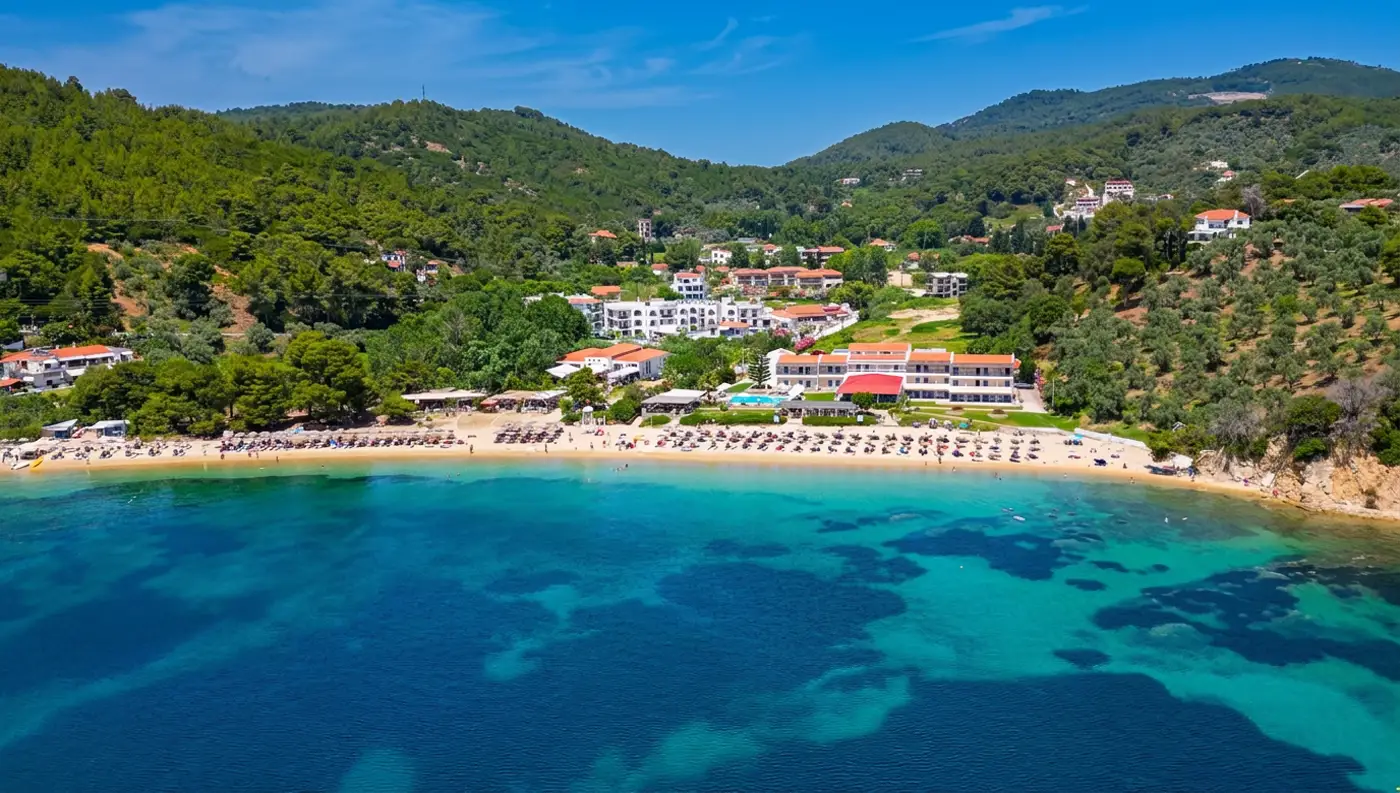 Εναέρια λήψη με τα κρυστάλλινα νερά, όπου ο Τρούλος Σκιάθου φαίνεται να περιβάλλεται από πυκνό πευκοδάσος.