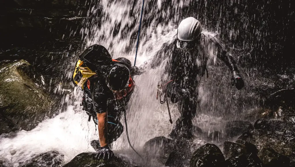 Δύο έμπειροι οδηγοί canyoning με κράνη και στολές νεοπρέν διασχίζουν ορμητικά νερά κάτω από καταρράκτη.
