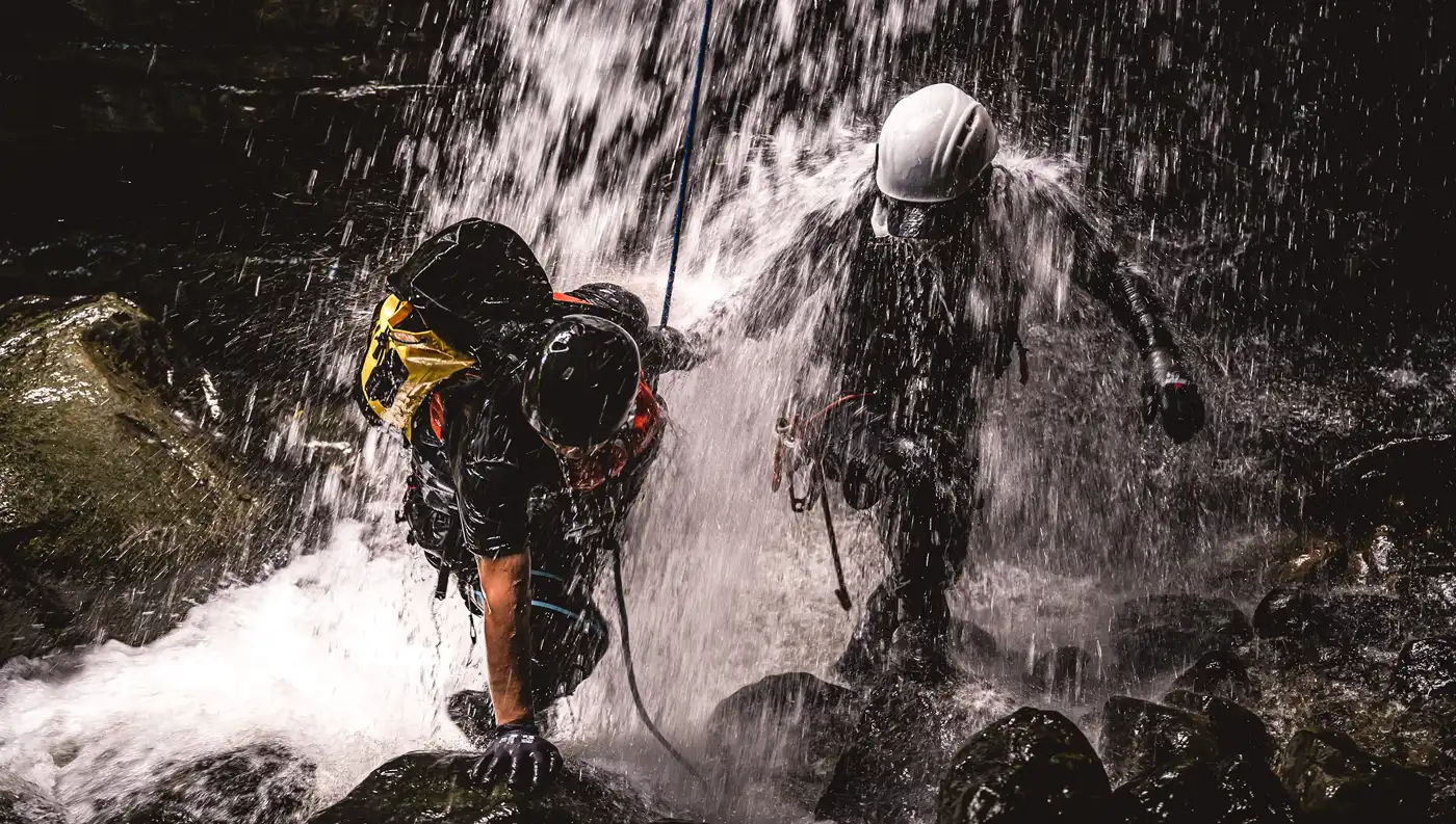 Δύο έμπειροι οδηγοί canyoning με κράνη και στολές νεοπρέν διασχίζουν ορμητικά νερά κάτω από καταρράκτη.