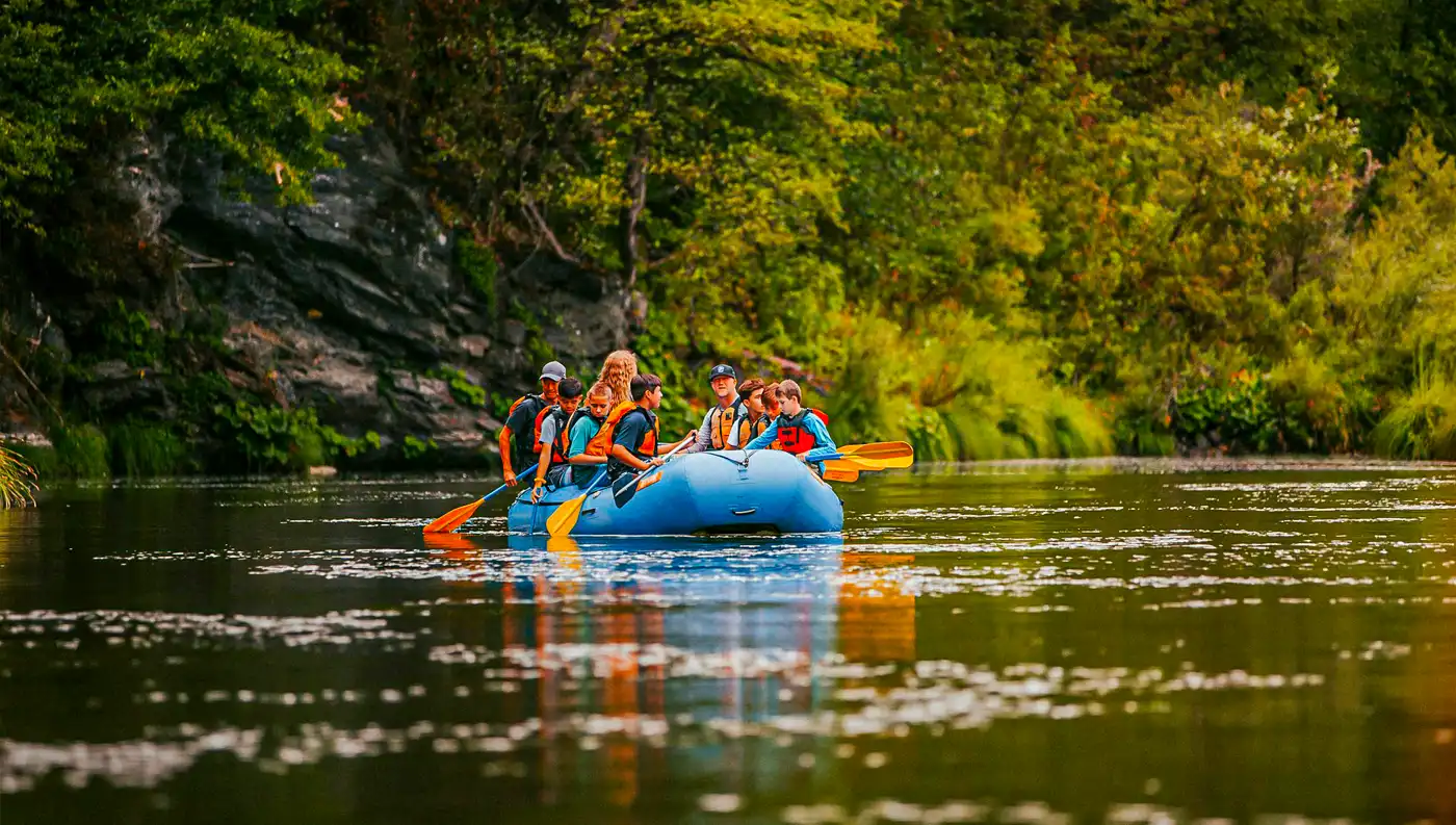 Οικογένεια και παιδιά σε μπλε βάρκα rafting σε ήρεμα, γαλήνια νερά ποταμού με δέντρα.