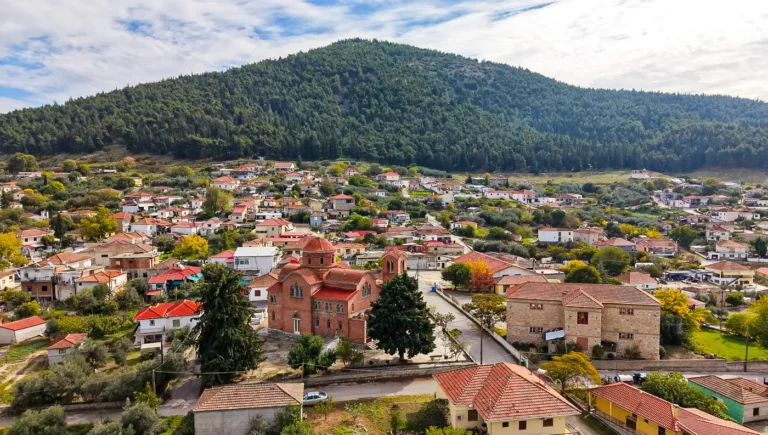 Αεροφωτογραφία από drone που δείχνει το χωριό Δομένικο Ελασσόνας απλωμένο αμφιθεατρικά, με τον Ιερό Ναό Παμμεγίστων Ταξιαρχών στο κέντρο και έναν μεγάλο κατάφυτο λόφο στο βάθος