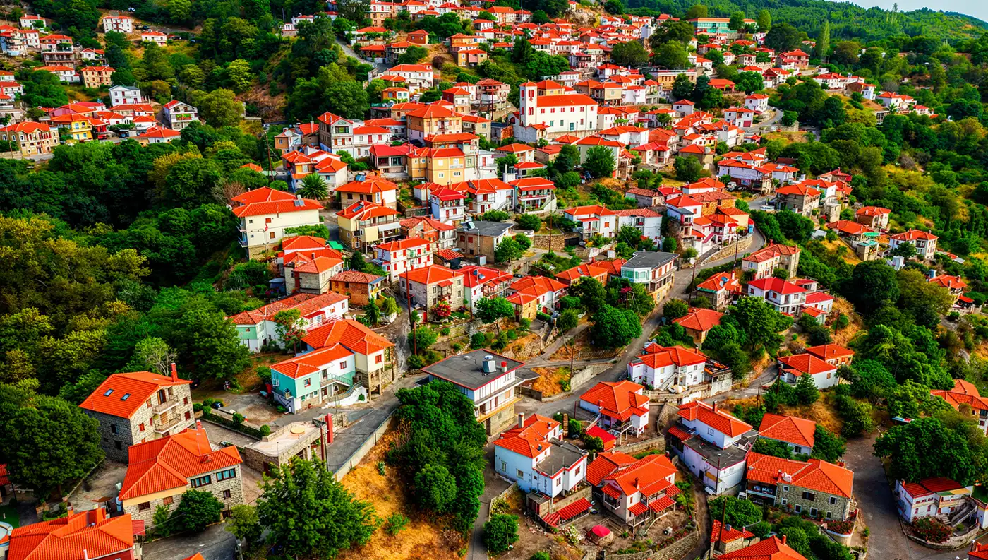 Εναέρια λήψη της Μελιβοίας, δείχνοντας τα σπίτια να απλώνονται στην καταπράσινη πλαγιά, στην αφετηρία για το Μονοπάτι Βελίκας.